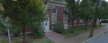 white doors to a brick building along a brick sidewalk with trees lining the street between the sidewalk and building.