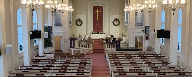 The interior of the Woodford's Church sanctuary from above. Lines of white pews and a rust red carpet aisle leading up to an alter with a gold cross hanging behind it, and two Christmas wreaths. 8 chandeliers hang from the slightly domed ceiling over the seating.